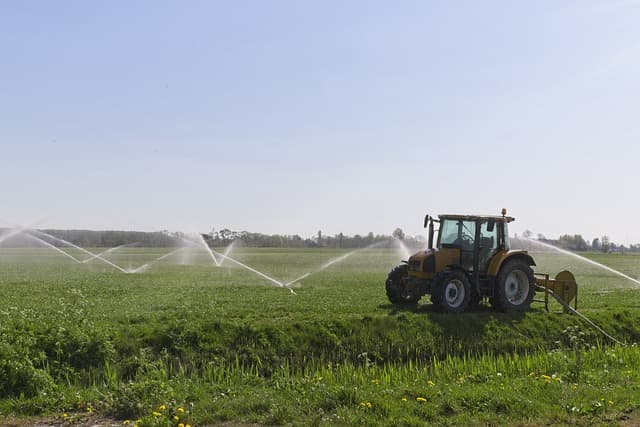 um campo de plantação bem verde com um trator a sua frente e aspersores jorrando água ao fundo